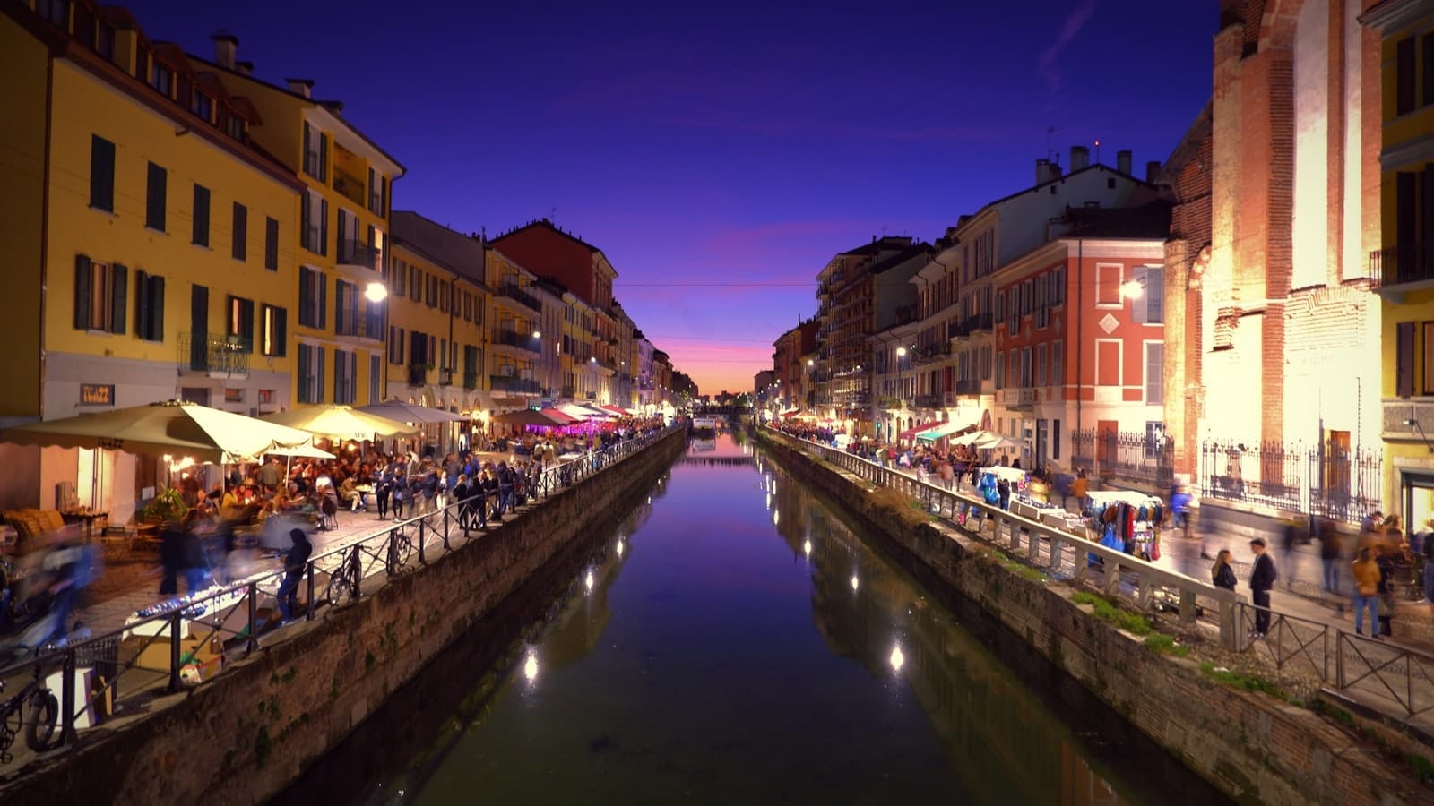 วิวยามเย็นริมคลอง Navigli ในมิลาน ไฟหลากสี ตึกสีสันสดใส เงาสะท้อนในน้ำ และผู้คนครึกครื้นริมคลองสองฝั่ง