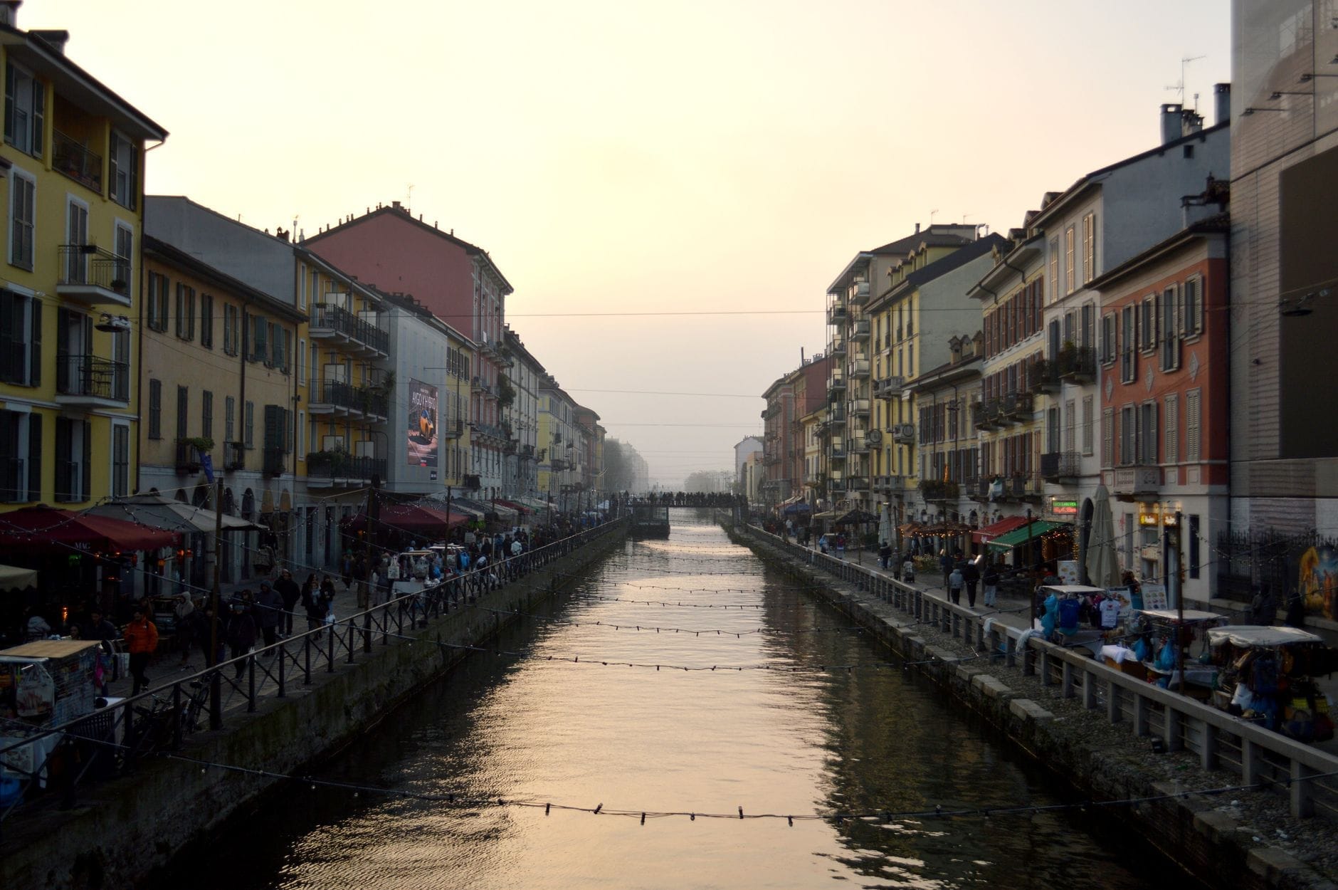 Vista serale lungo il Naviglio Grande di Milano con gli edifici storici dai colori vivaci, la folla animata e i riflessi sull'acqua sotto un cielo acceso dal tramonto.