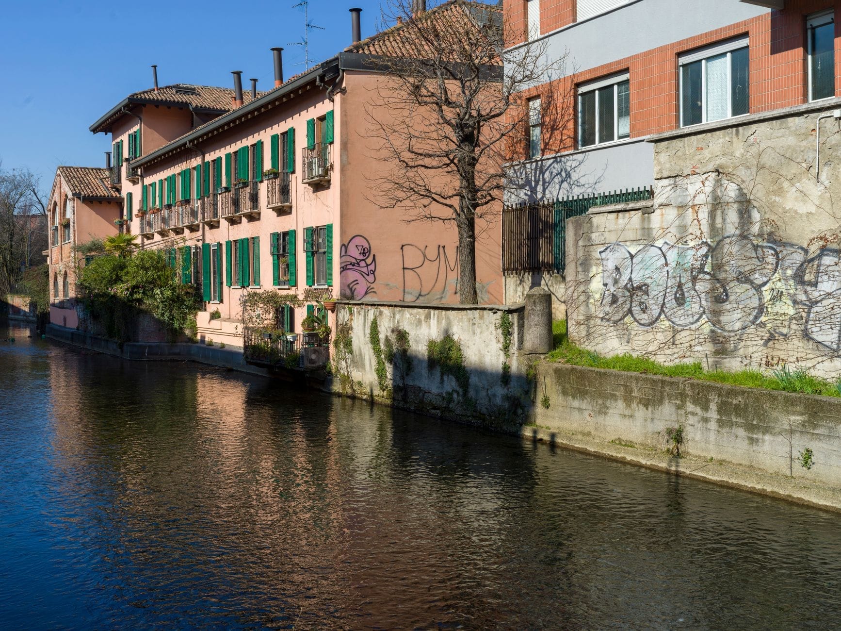 Scena tranquilla sul canale con i tipici edifici rosa e in mattoni dai battenti verdi, il riflesso degli alberi nell'acqua e l'atmosfera serena lungo il Naviglio Pavese a Milano.