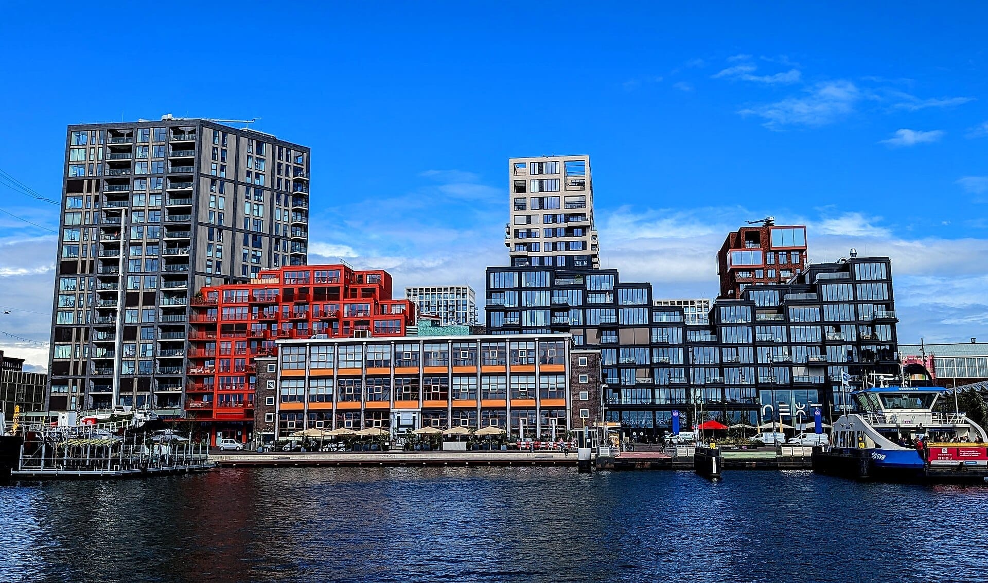 Edificios geométricos modernos del NDSM Wharf se reflejan en el río IJ bajo un cielo azul brillante con un ferry amarrado en el muelle.