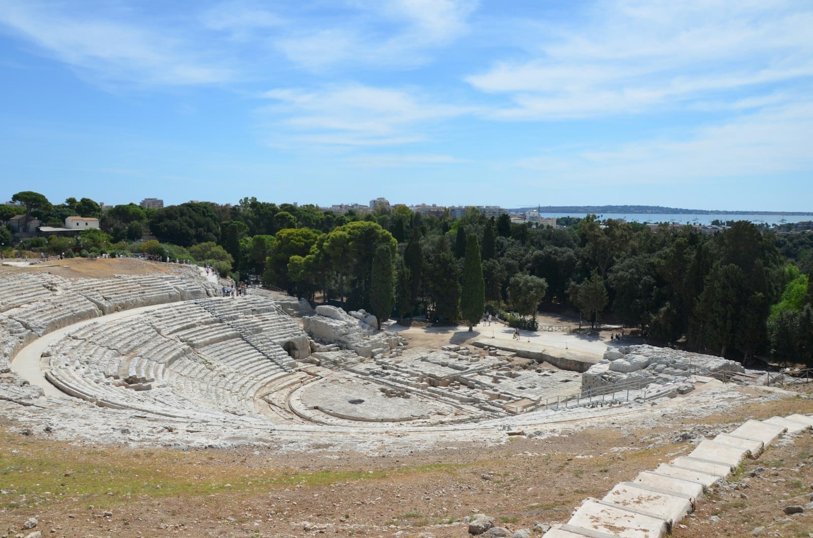 Weitwinkelblick auf das antike griechische Theater im Archäologischen Park Neapolis in Syrakus, mit steinernen Sitzreihen, üppiger Vegetation und weitem Himmel.