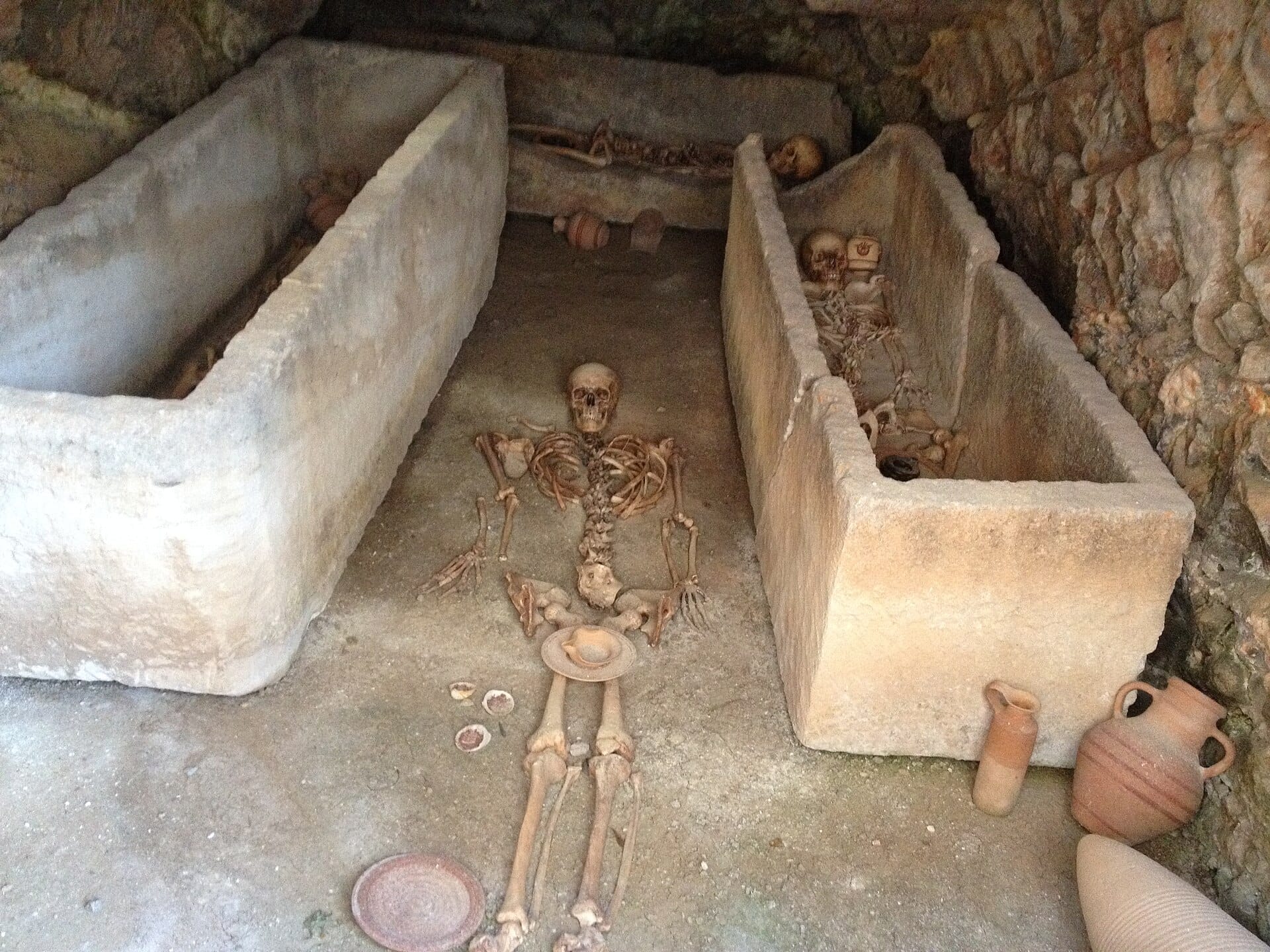 Antiche tombe di pietra con scheletri umani e ceramiche alla Necropolis del Puig des Molins a Ibiza, circondate da rozze pareti di pietra.