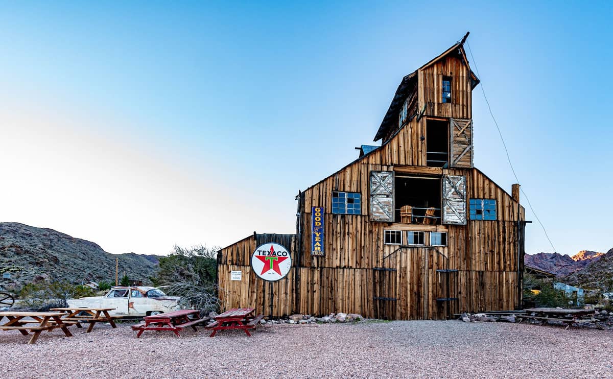 Bangunan tambang kayu tua besar dengan papan TEXACO, meja piknik, dan perbukitan gurun di Nelson Ghost Town, Eldorado Canyon, Nevada.