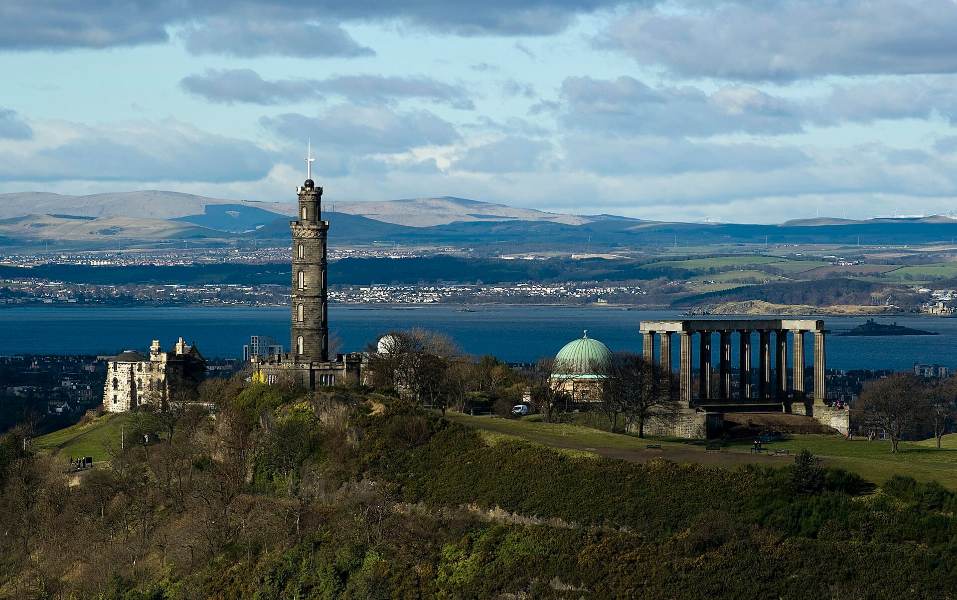 Das Nelson Monument thront markant auf dem Calton Hill und überblickt Edinburgh mit Blick auf das Meer und die umliegenden Sehenswürdigkeiten unter blauem Himmel.