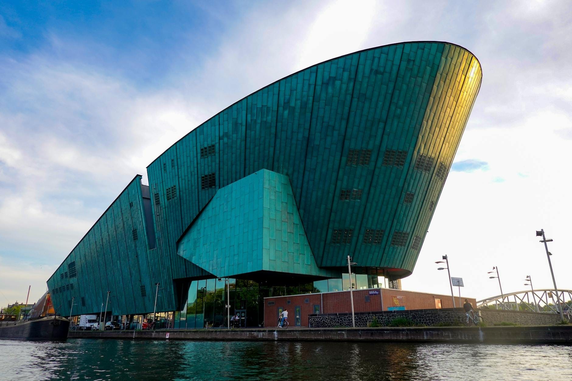 El llamativo Museo de Ciencias NEMO, revestido de cobre verde, visto desde el otro lado del paseo marítimo, con cielo azul y personas caminando cerca de la entrada.