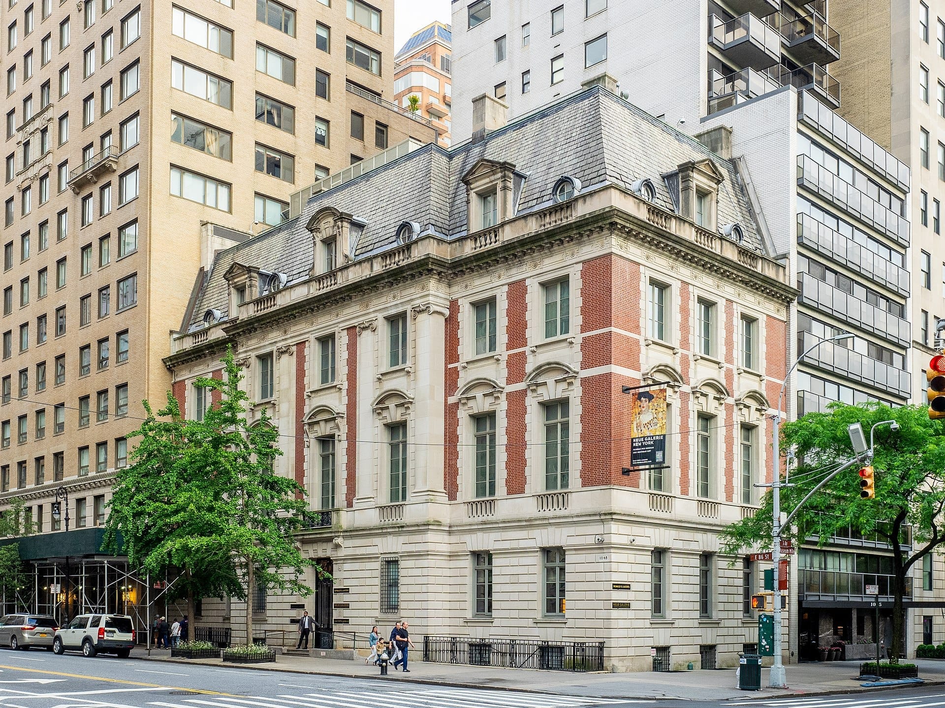 Vista esterna della Neue Galerie New York, uno storico palazzo in stile Beaux-Arts con facciata in mattoni rossi e pietra all'angolo di una strada cittadina.