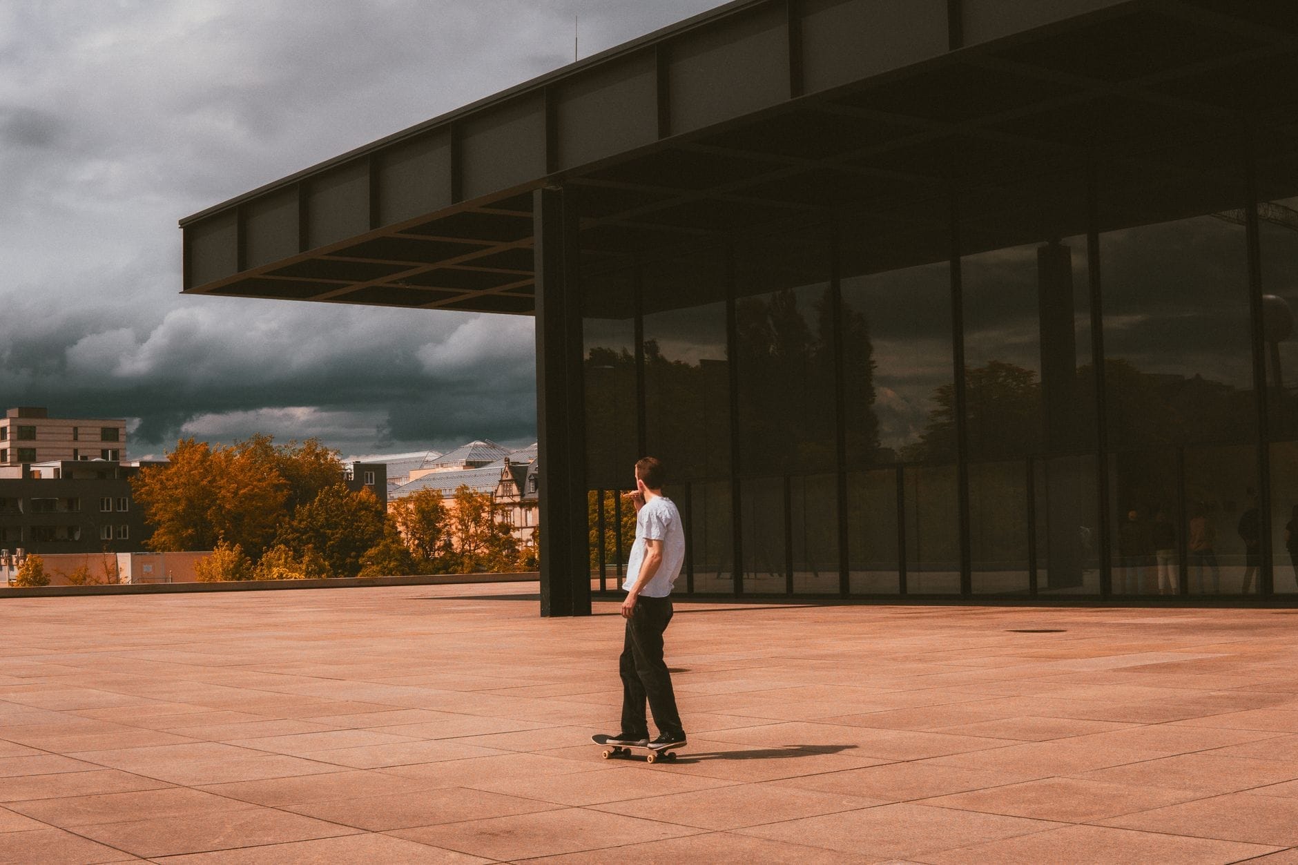 Una persona sullo skateboard davanti alla Neue Nationalgalerie, con la spettacolare facciata in vetro e la pensilina in acciaio stagliate su un cielo drammatico.