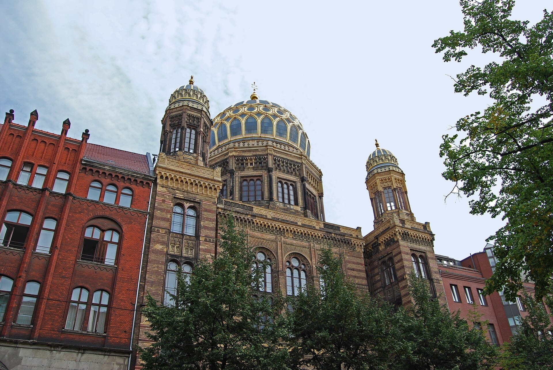 La Nuova Sinagoga di Berlino presenta elaborate cupole dorate e un'architettura di ispirazione moresca, incorniciata da edifici in mattoni rossi e alberi verdi frondosi sotto un cielo luminoso.