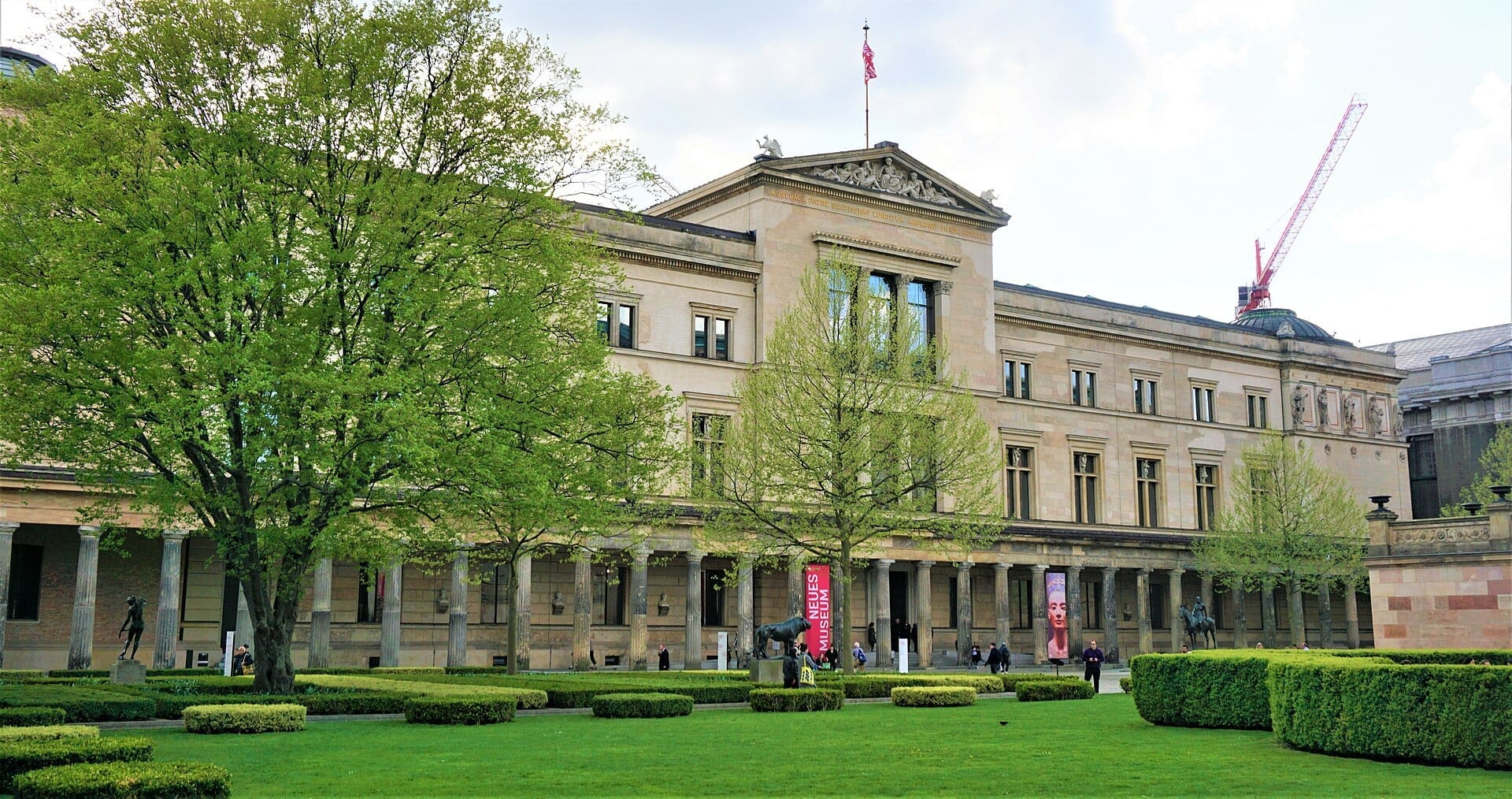 L'esterno del Neues Museum a Berlino, incorniciato da alberi verdi e giardini, con visitatori che camminano vicino alla grandiosa facciata neoclassica.
