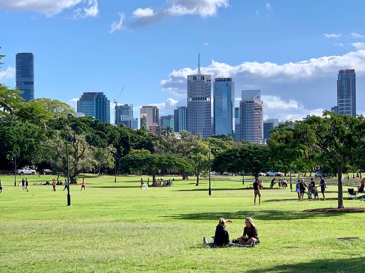 Weitläufige grüne Rasenflächen und vereinzelte Bäume im New Farm Park mit Menschen, die sich entspannen und spielen, vor der modernen Skyline von Brisbane unter blauem Himmel.