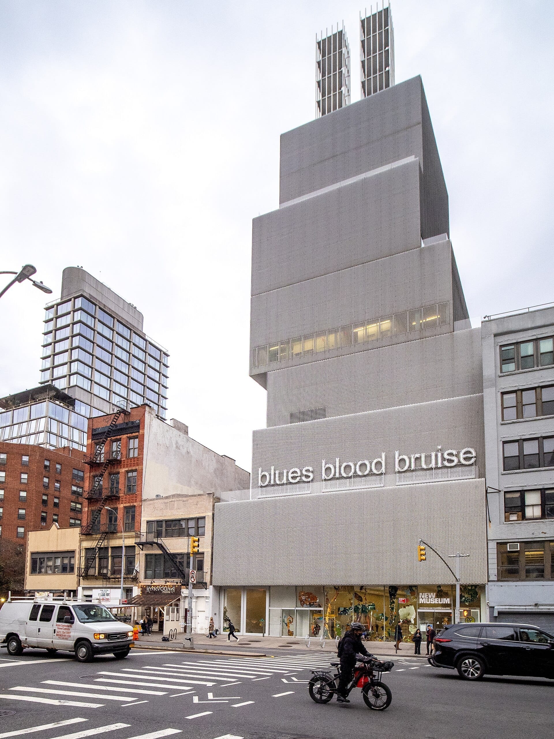 La caratteristica facciata grigia sovrapposta del New Museum domina una trafficata strada del Lower East Side, con persone e veicoli che transitano davanti al crepuscolo.