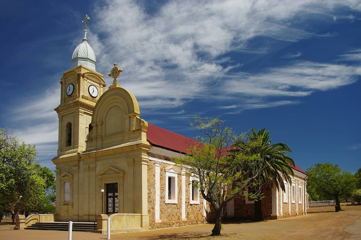 Die historische Steinkirche in New Norcia steht unter einem lebhaften blauen Himmel, umgeben von grünen Bäumen und auffälligen Wolkenformationen.