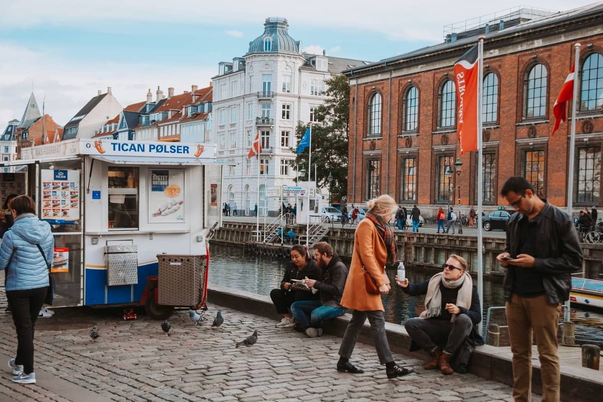 Street food cart by a canal in Copenhagen, with people sitting and enjoying food along the waterfront, historic buildings visible in background.