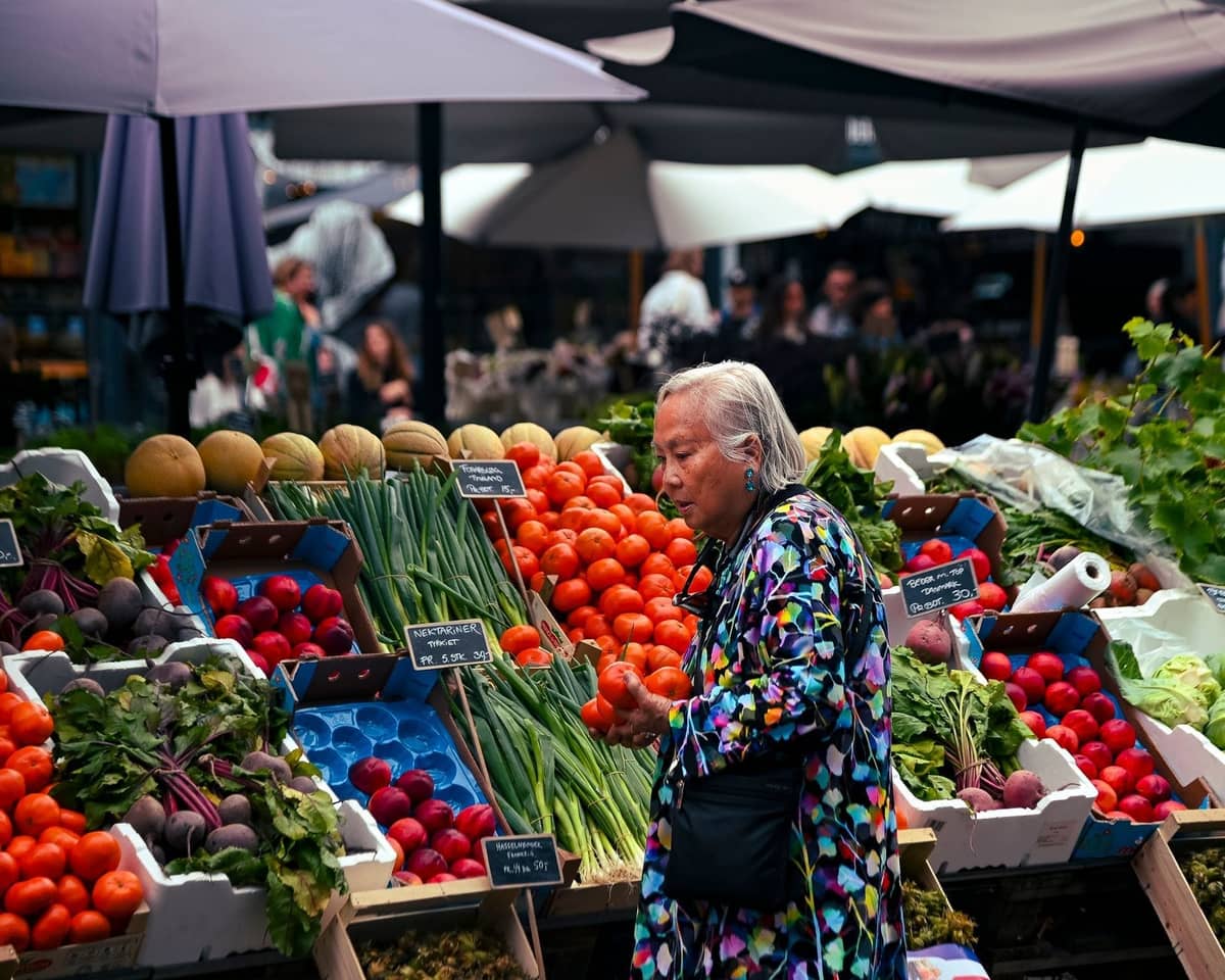 Woman shopping at an outdoor Copenhagen market stall with a colorful display of seasonal vegetables and fruits.