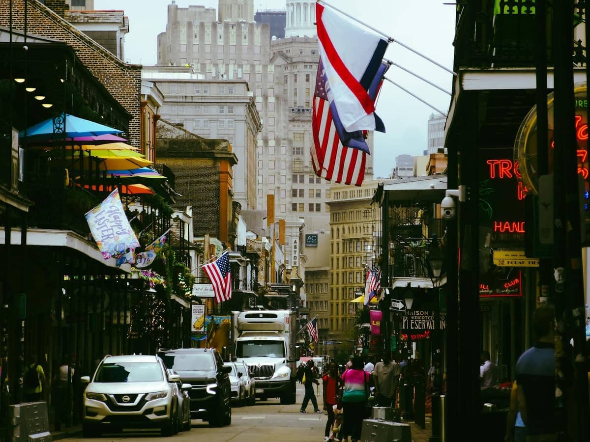 A busy and colorful street scene on Bourbon Street in New Orleans, with flags, tourists, and storefronts lining the avenue.