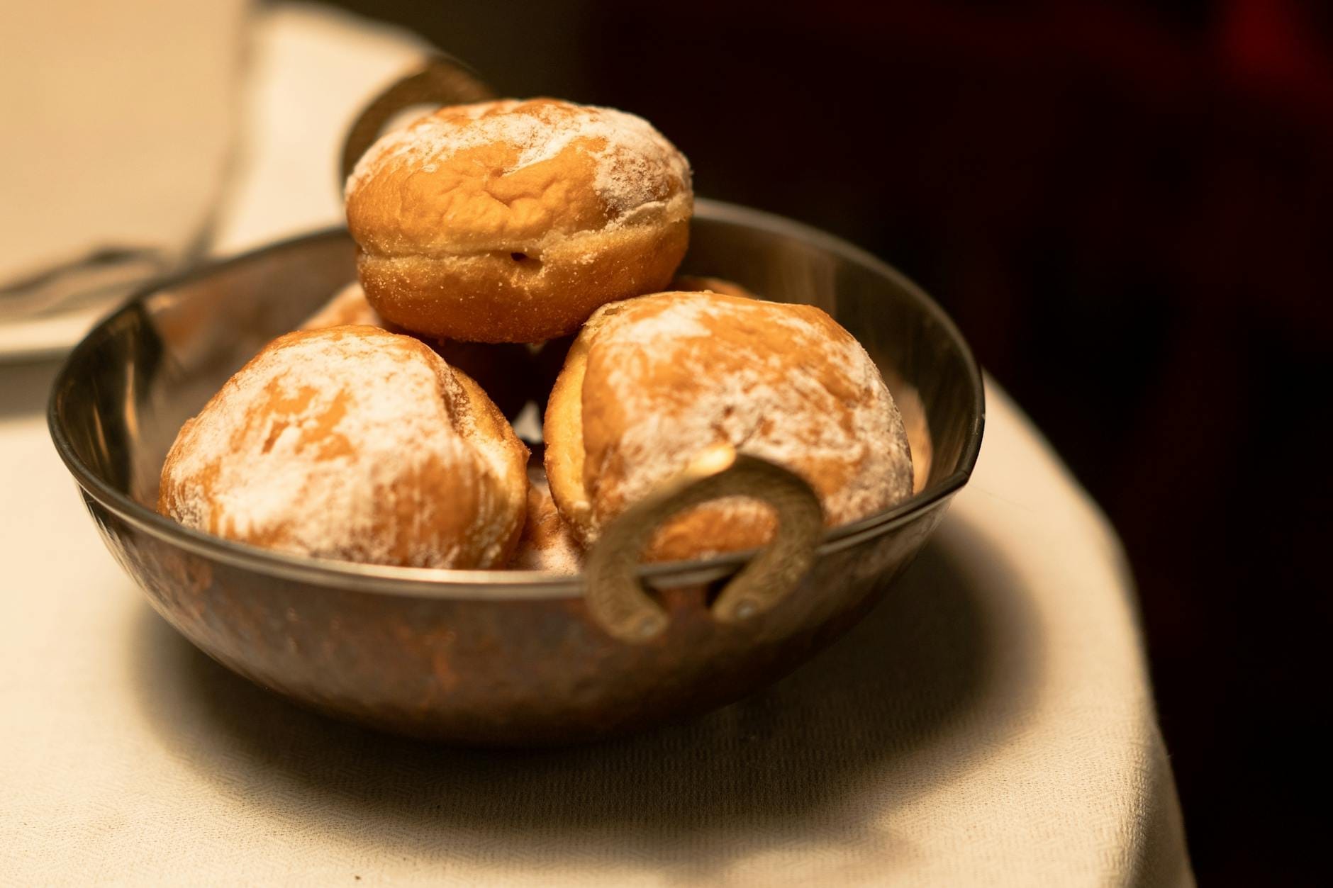 Close-up de beignets dourados polvilhados com açúcar em pó em uma tigela de metal sobre uma toalha de mesa branca, com iluminação suave em ambiente interno.