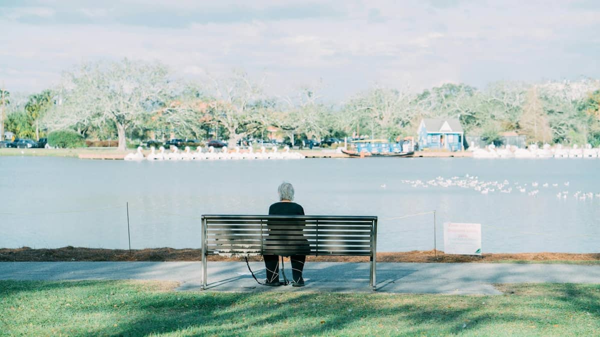An older person sits alone on a bench facing the lagoon in New Orleans City Park, with trees and a blue boathouse in view.
