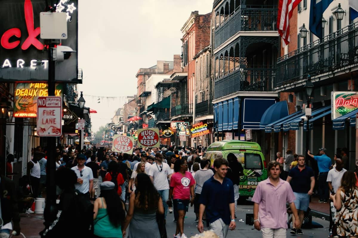 A lively New Orleans street scene with crowds walking among historic French Quarter buildings and neon signs.