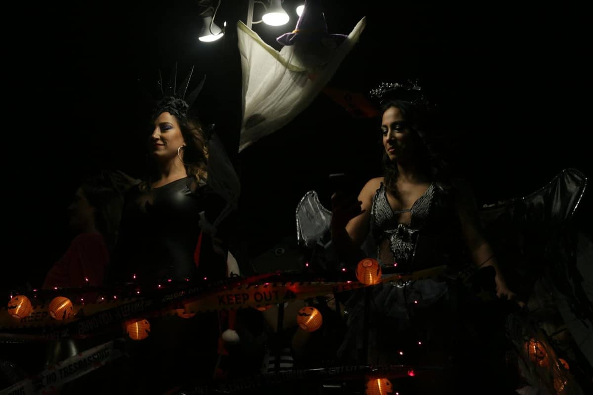 Two women in elaborate black costumes riding a float decorated with Halloween lights and caution tape at night.