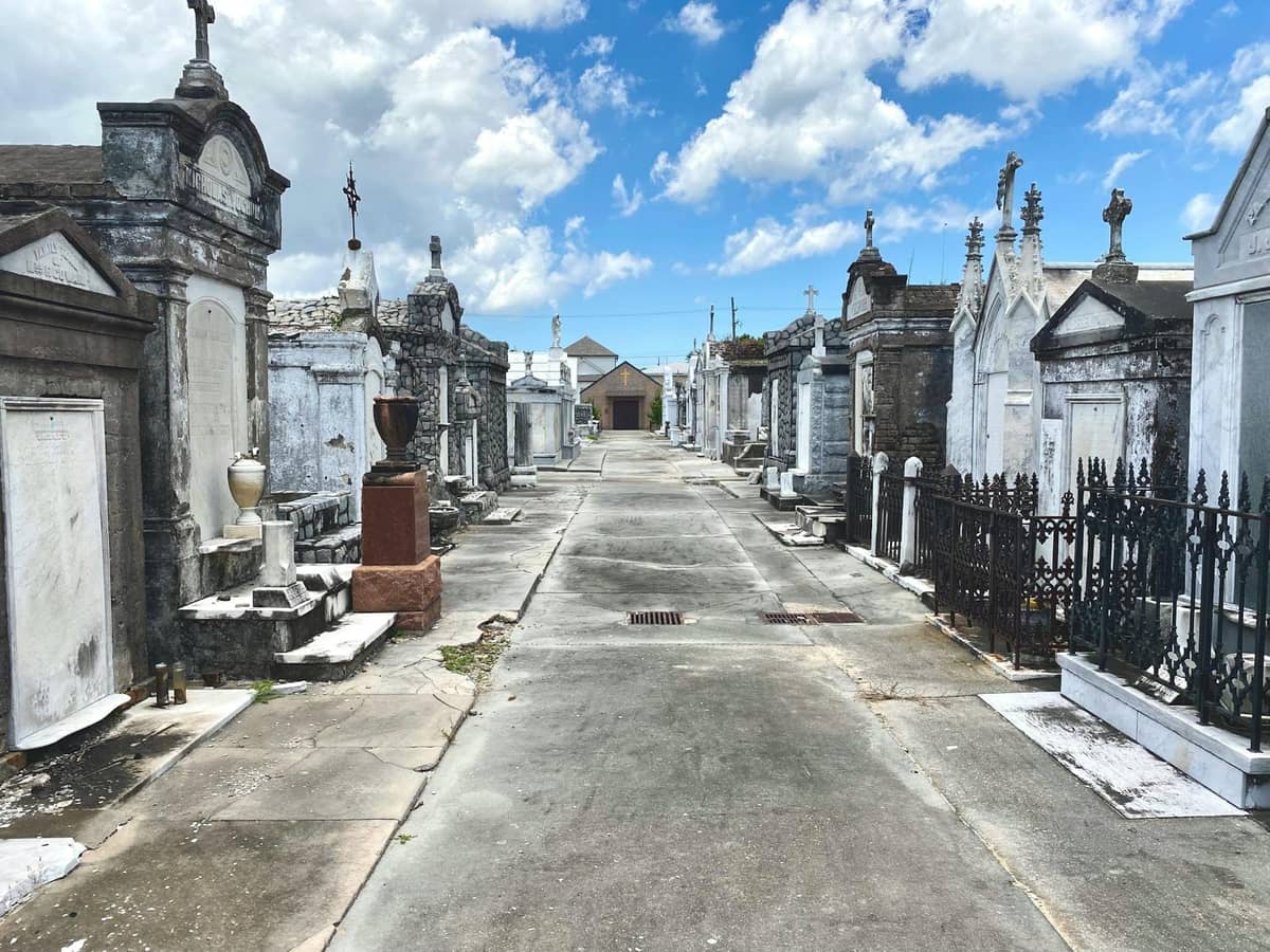 Above-ground tombs line a pathway in an historic cemetery under a bright blue sky with scattered clouds.