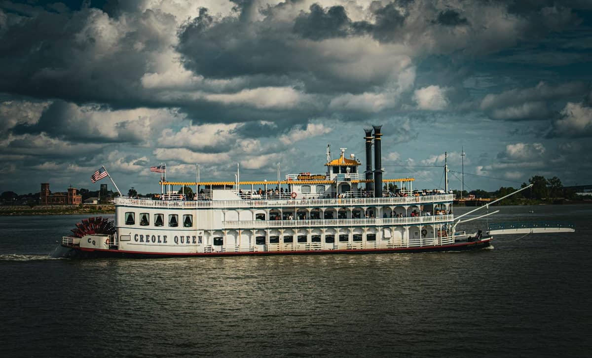 A classic white river steamboat named Creole Queen sails on the Mississippi River under a dramatic cloudy sky.