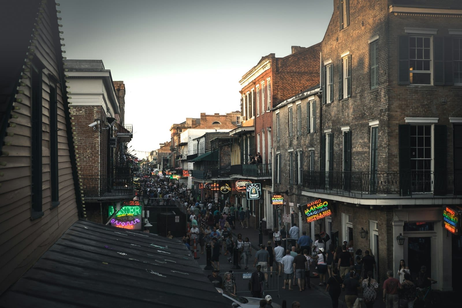 Multidões caminham pela Bourbon Street no French Quarter de Nova Orleans, rodeadas por edifícios históricos de tijolo e letreiros de néon brilhantes ao entardecer.