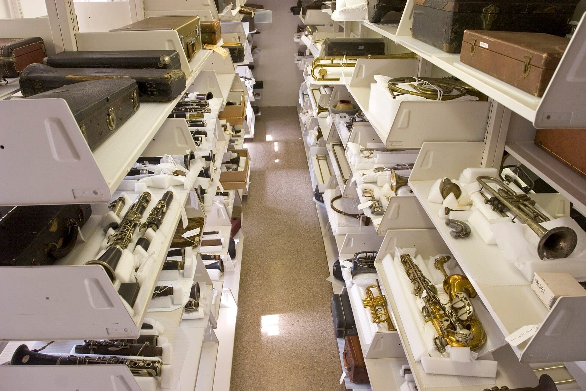 File di scaffali bianchi al New Orleans Jazz Museum riempiti di strumenti a fiato vintage in ottone, clarinetti e vecchie custodie per strumenti, sotto illuminazione fluorescente.
