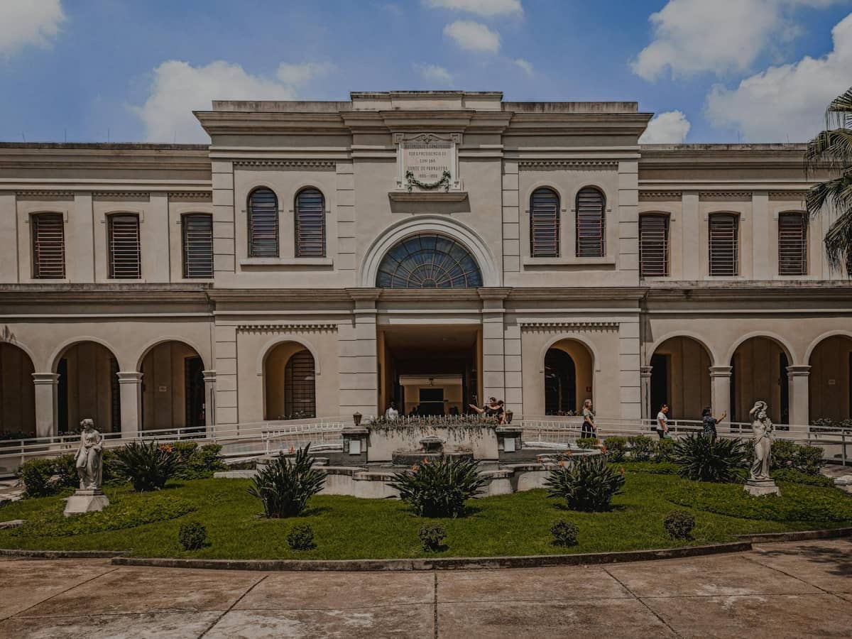 Elegant stone building with arched windows and a central courtyard featuring statues and green landscaping under a blue sky.