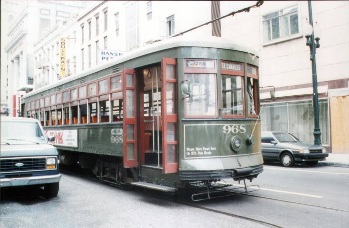 Historic green St. Charles streetcar on a New Orleans avenue, with city buildings and cars visible, referencing the Mardi Gras parade route.