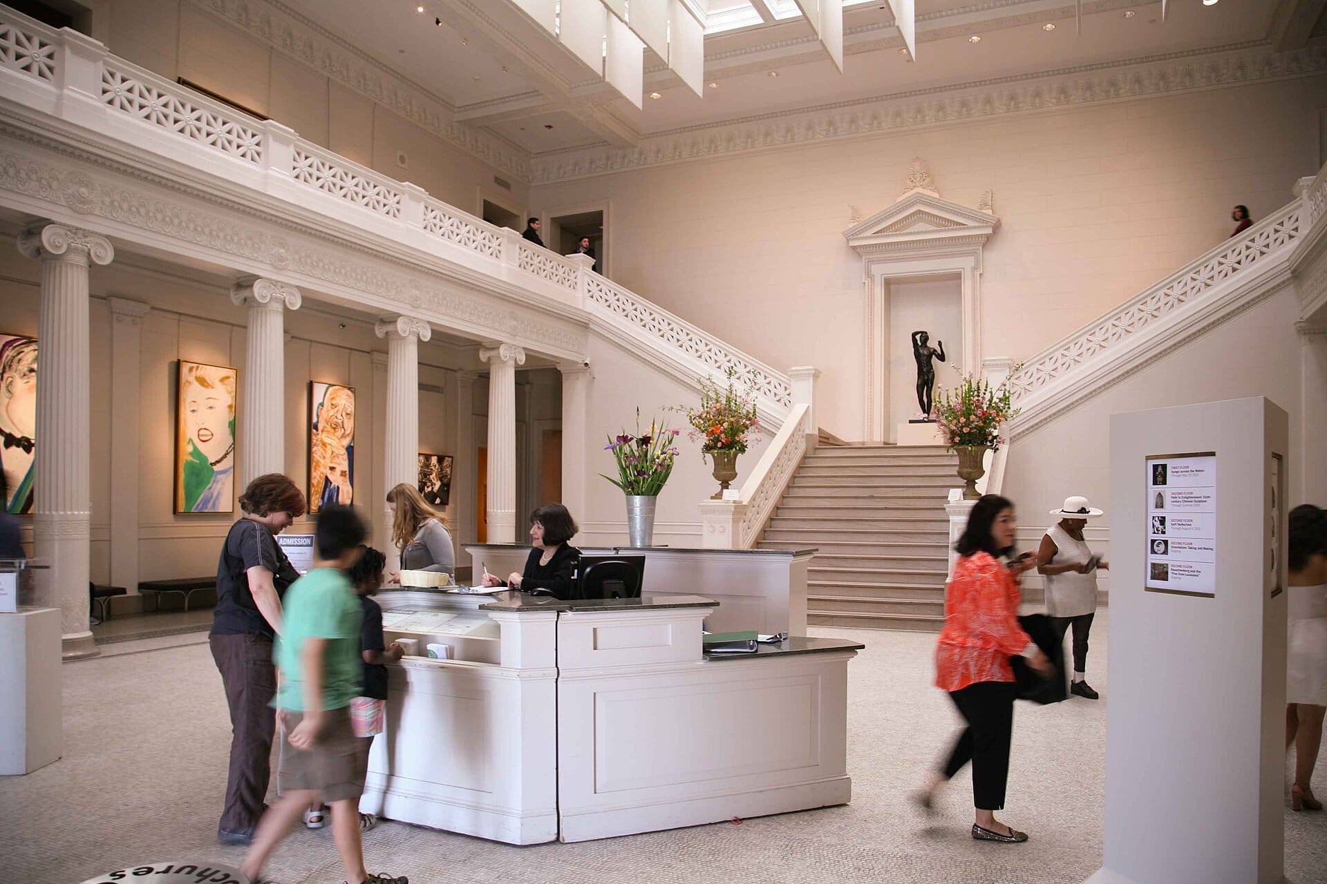 Il grande atrio d'ingresso del New Orleans Museum of Art con visitatori, un banco della reception, una scala in marmo e dettagli architettonici classici.