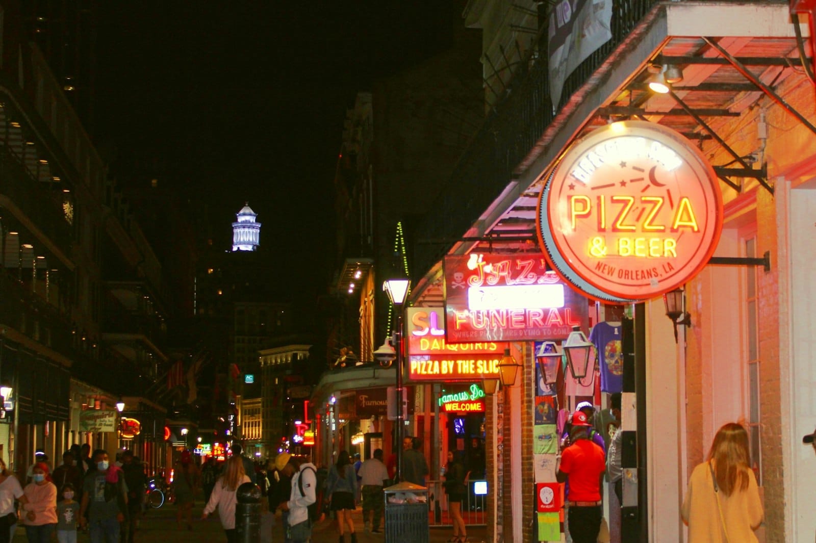 Pessoas caminham por uma movimentada rua de New Orleans à noite, iluminada por letreiros de néon de bares e pizzarias e luzes coloridas da cidade.