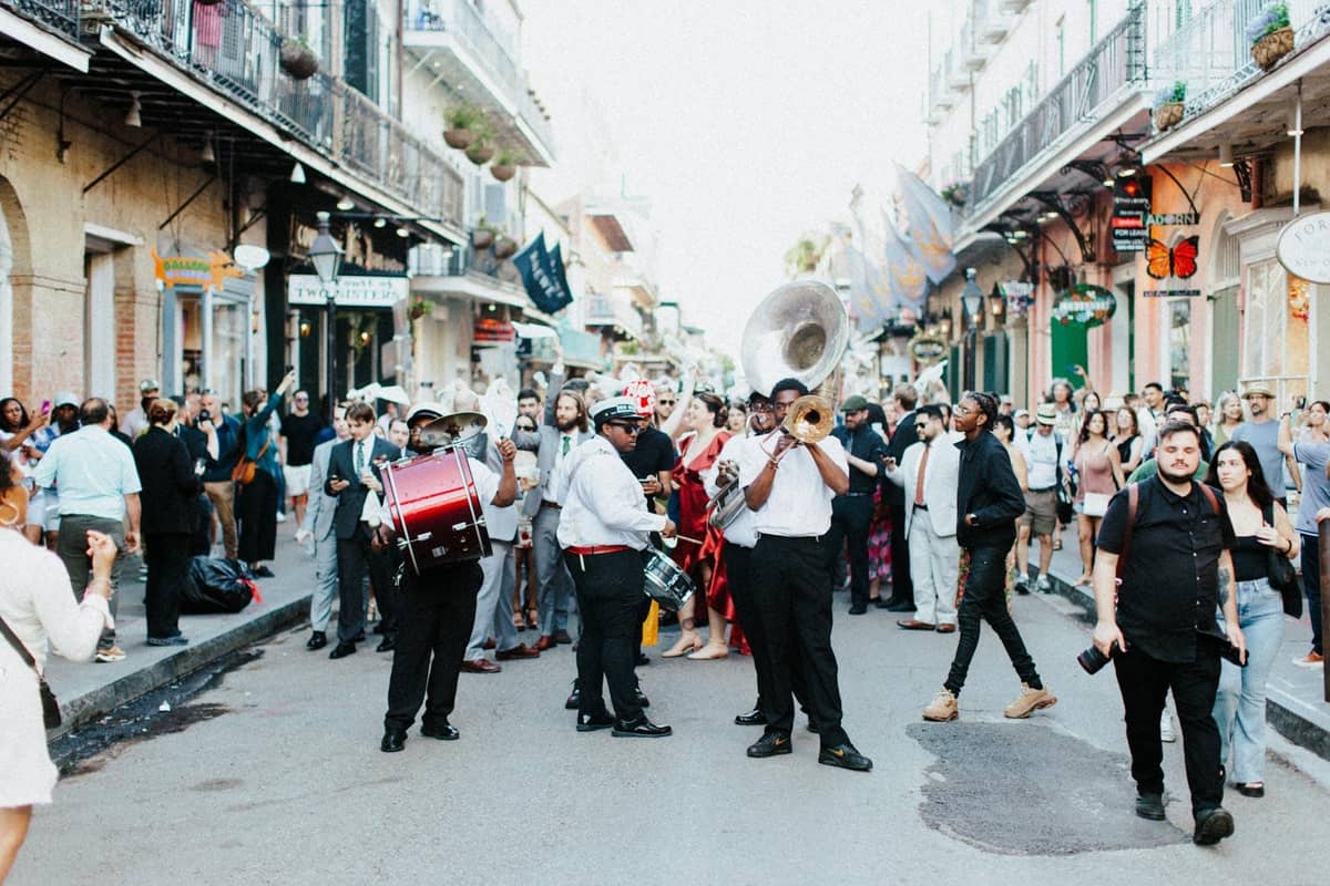 A brass band leads a lively crowd through a historic New Orleans street lined with balconies and local shops during a daytime parade.