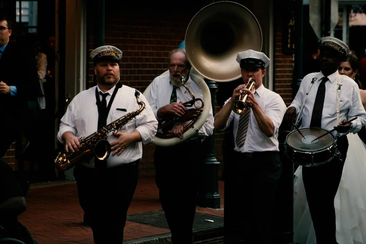 A brass band marches on a New Orleans street with musicians playing saxophone, sousaphone, trumpet, and snare drum, wearing white shirts and band hats.
