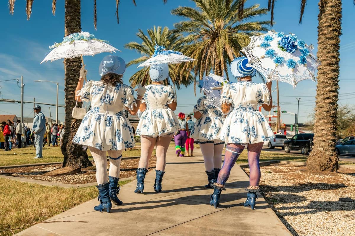 A group of women in coordinated blue and white costumes holding decorated parasols walk in a festive parade with palm trees and bystanders in New Orleans.