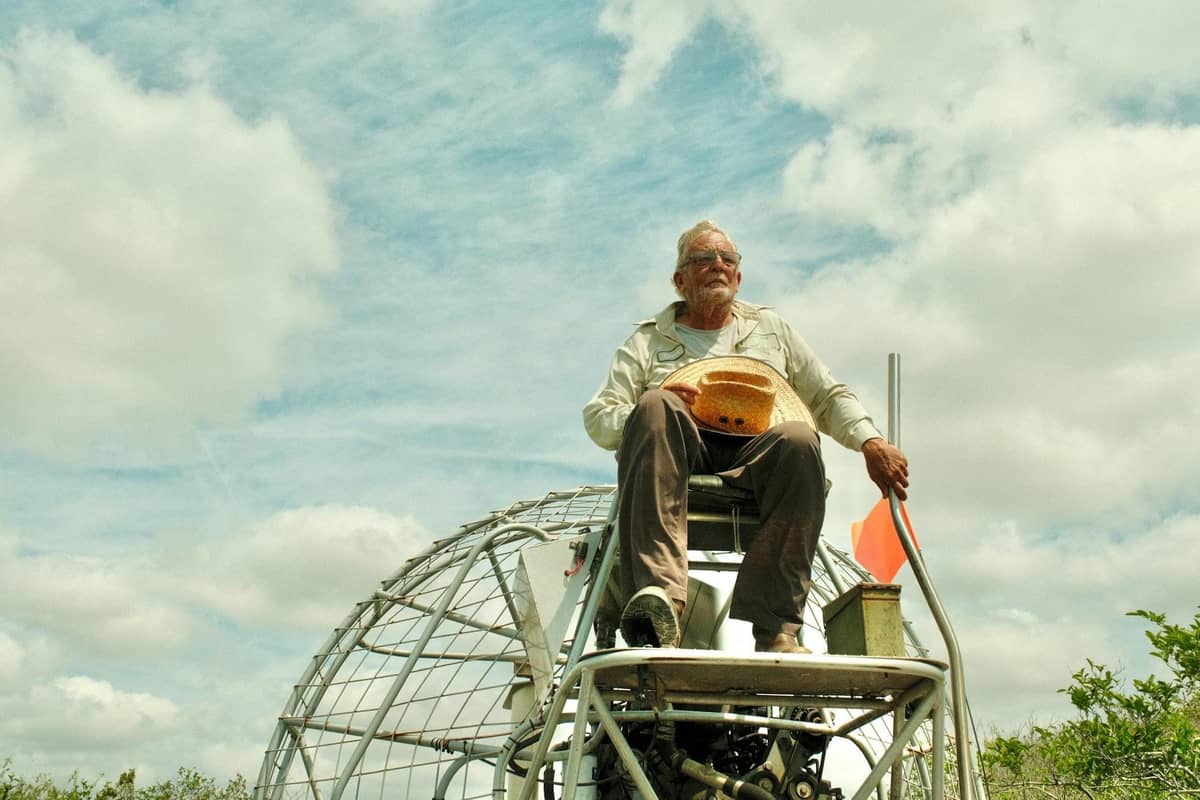 Man sitting atop an airboat with fan and cage visible, surrounded by marshland under a blue sky.