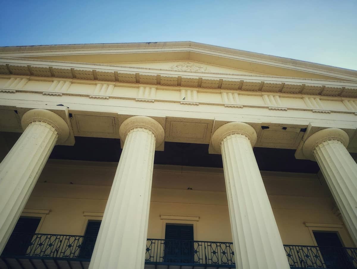 Low angle view of a historic Greek Revival building with large columns and ironwork balcony under a blue sky.