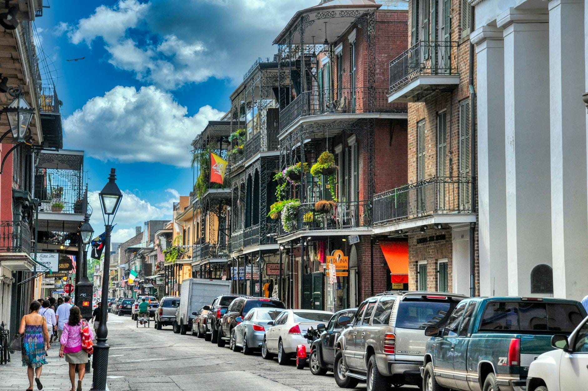 Uma rua movimentada de Nova Orleans com pessoas caminhando, prédios históricos com varandas de ferro forjado e plantas penduradas, sob um céu azul com nuvens.