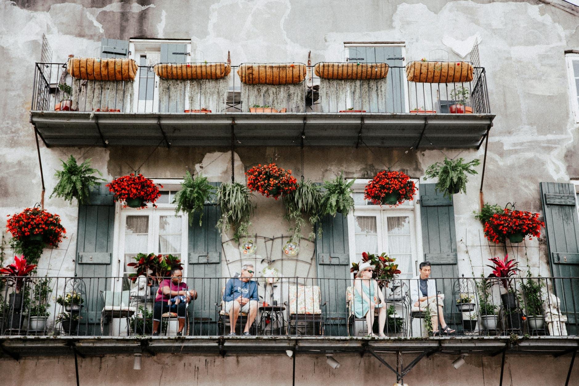 Famílias e crianças reunidas em uma varanda clássica do French Quarter de Nova Orleans, decorada com flores e plantas coloridas, transmitindo uma atmosfera acolhedora e animada para toda a família.