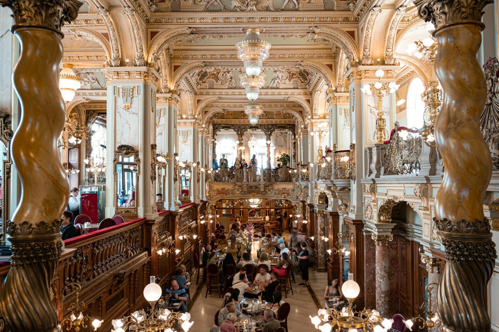 Interno sontuoso del New York Café a Budapest: colonne di marmo, soffitti dorati, lampadari e ospiti a tavola nella sala principale.