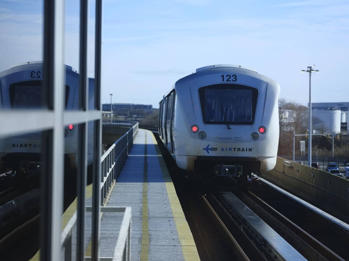 JFK AirTrain กำลังเข้าสู่ชานชาลาสถานียกระดับ มองเห็นรางรถไฟ แสงกลางวันสว่างจ้า บรรยากาศระบบขนส่งสมัยใหม่