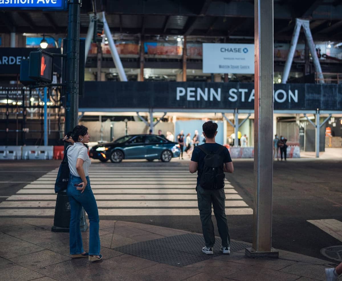สองคนรอข้ามถนนที่ทางม้าลายหน้า Penn Station พร้อมป้ายที่มองเห็นได้ชัดและบรรยากาศถนนในเมืองยามค่ำคืน