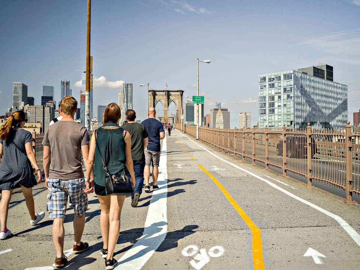 ผู้คนเดินบนทางเดินสำหรับคนเดินเท้าของ Brooklyn Bridge โดยมีตึกระฟ้าของ Manhattan เป็นฉากหลังในวันที่แดดจัด