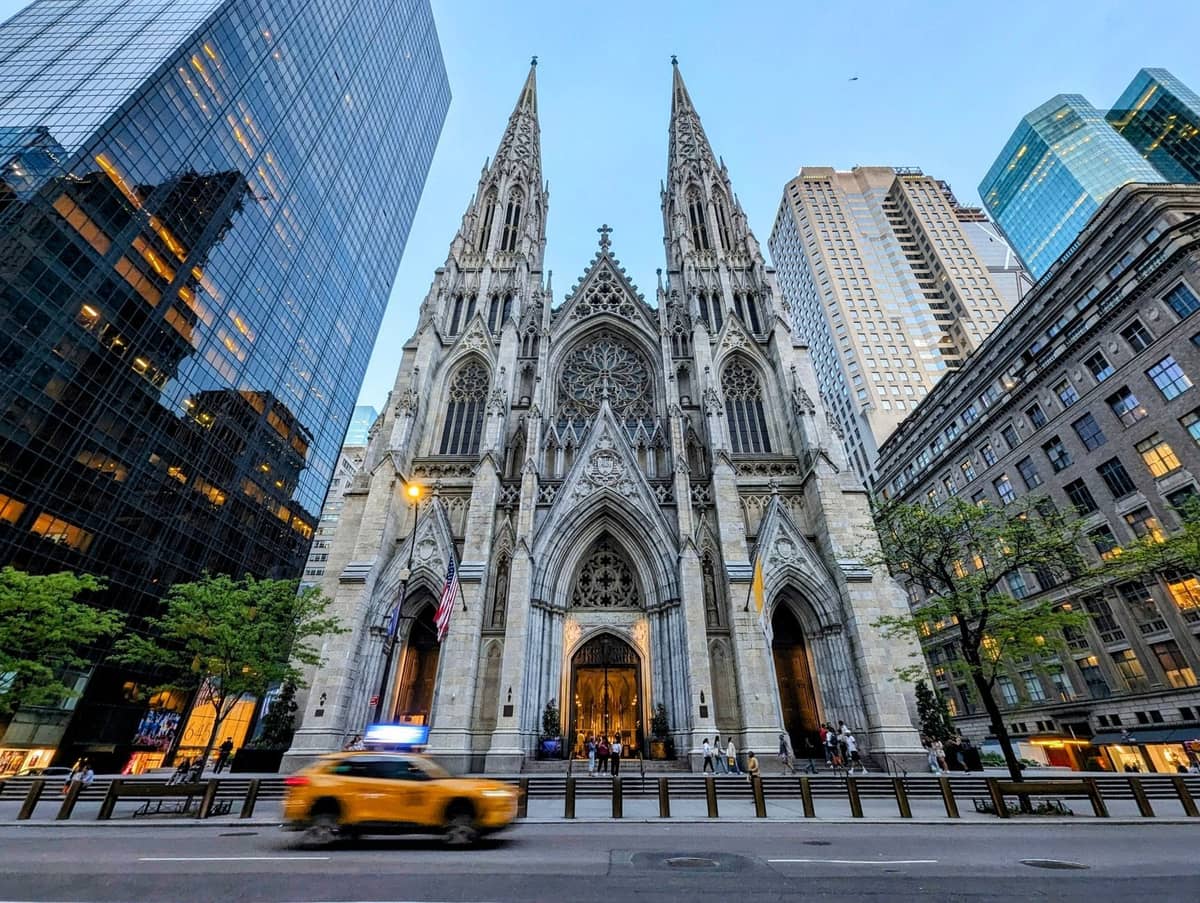 วิวกว้างของ St. Patrick's Cathedral พร้อมด้านหน้าแบบโกธิก ล้อมรอบด้วยตึกระฟ้าสมัยใหม่และแท็กซี่สีเหลืองของ NYC ในเบื้องหน้า
