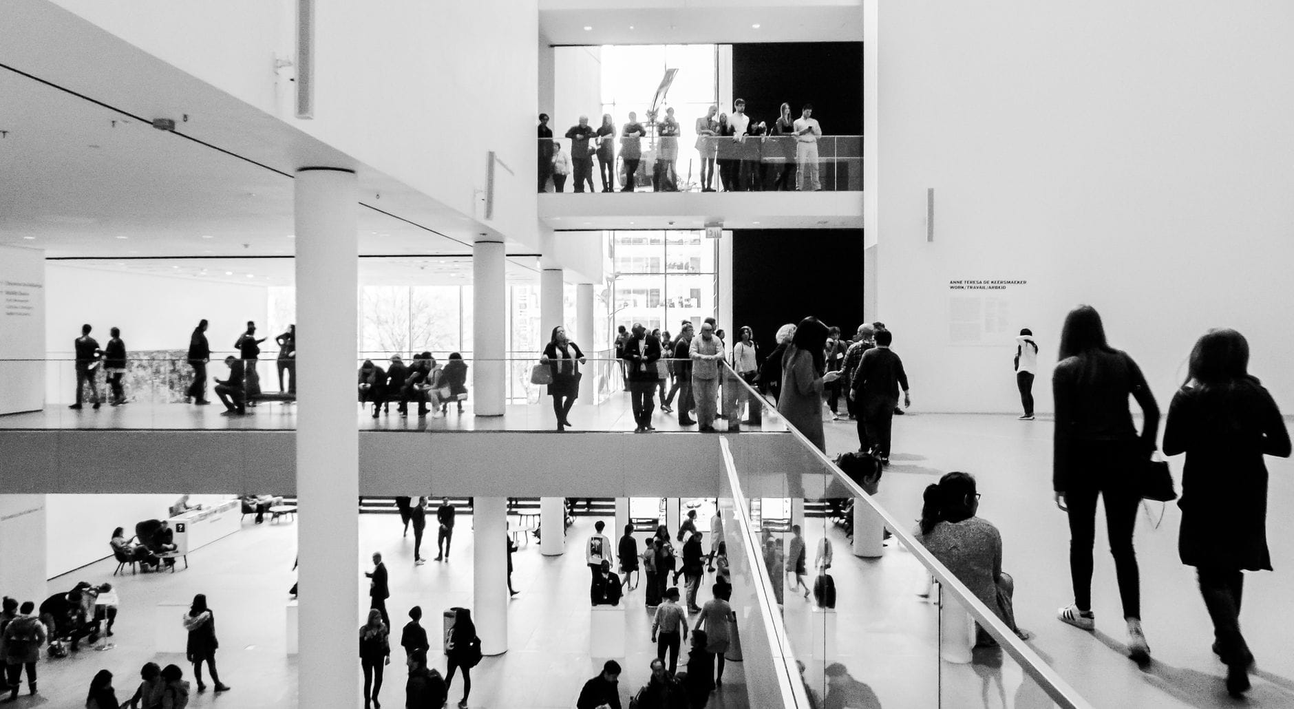 Interior em preto e branco de um museu de arte moderna com pessoas circulando e explorando múltiplos andares e espaços abertos banhados de luz natural.