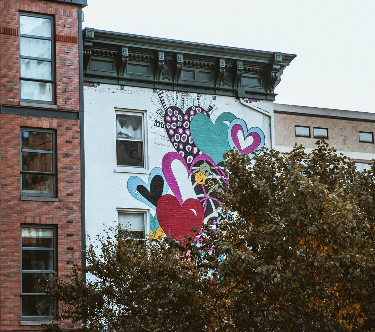 Buntes Street-Art-Wandgemälde mit aufgemalten Herzen an der Seite eines Gebäudes in New York City, teilweise von Ästen verdeckt.