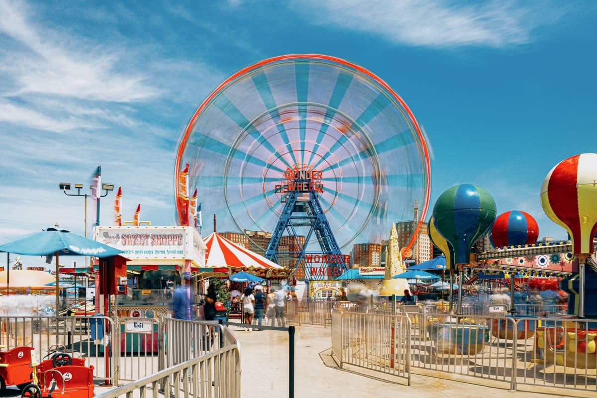 Helle Aussicht auf den Freizeitpark Coney Island mit dem Wonder Wheel, Karussells, bunten Buden und Menschen, die die Strandpromenade unter einem blauen Himmel genießen.