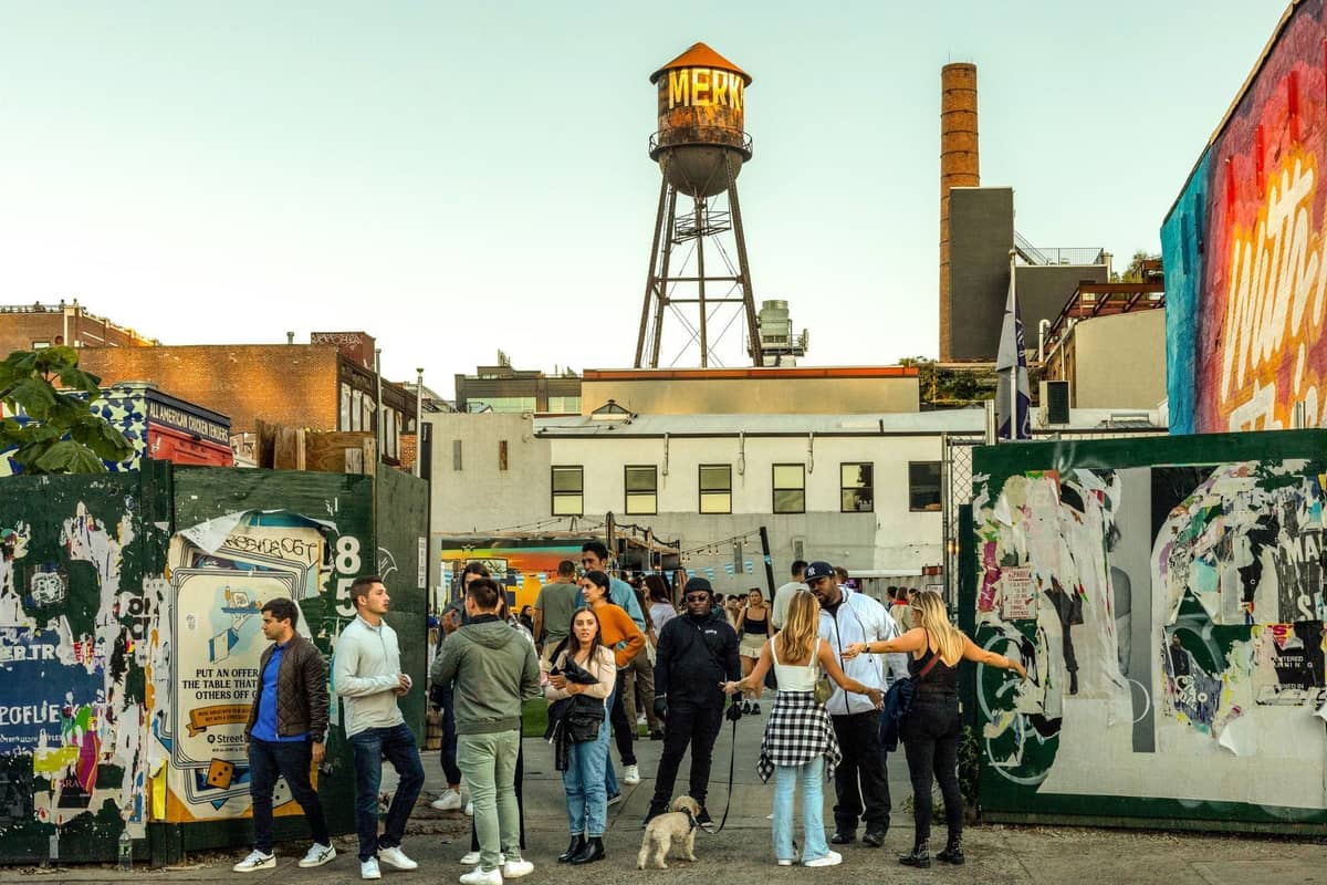 Gruppe junger Menschen an bunten Marktständen in Williamsburg mit dem markanten Wasserturm und Street-Art im Hintergrund.