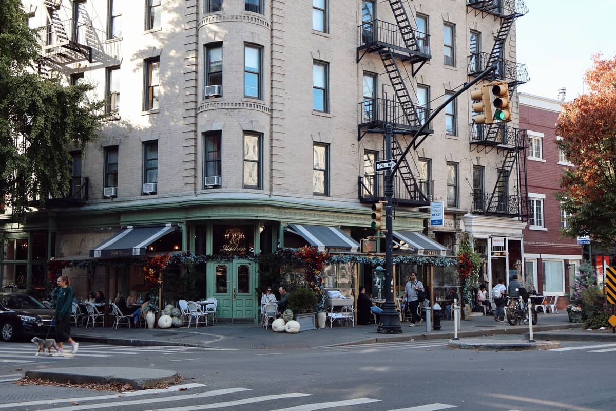 Eckansicht einer Straße in Greenwich Village mit einem klassischen New Yorker Gebäude, einem Café mit grüner Markise, im Freien speisenden Menschen und ikonischen Feuerleitern.