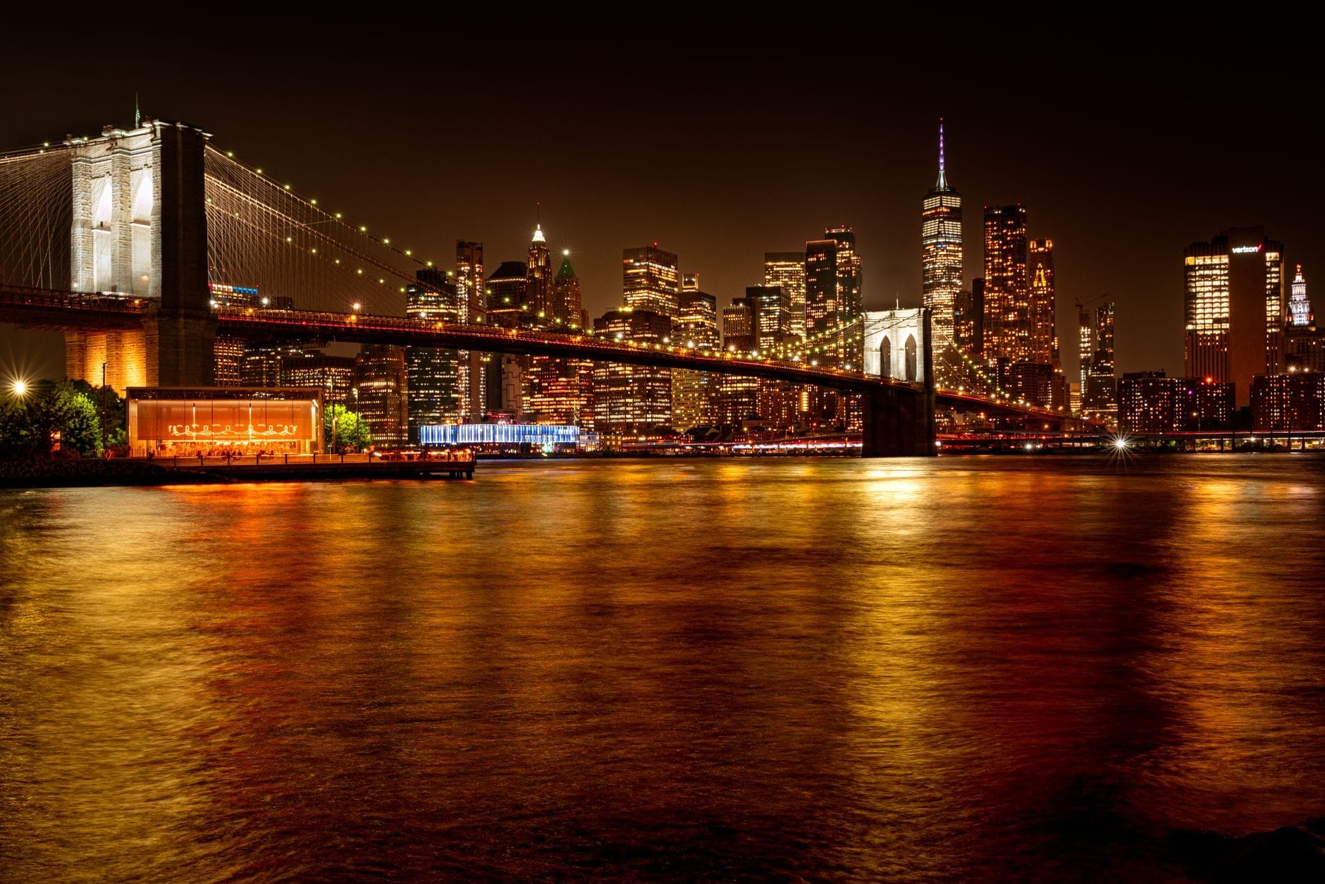 Vista noturna da Ponte do Brooklyn e do skyline de Nova York refletidos na água, com luzes brilhantes e uma atmosfera urbana vibrante.