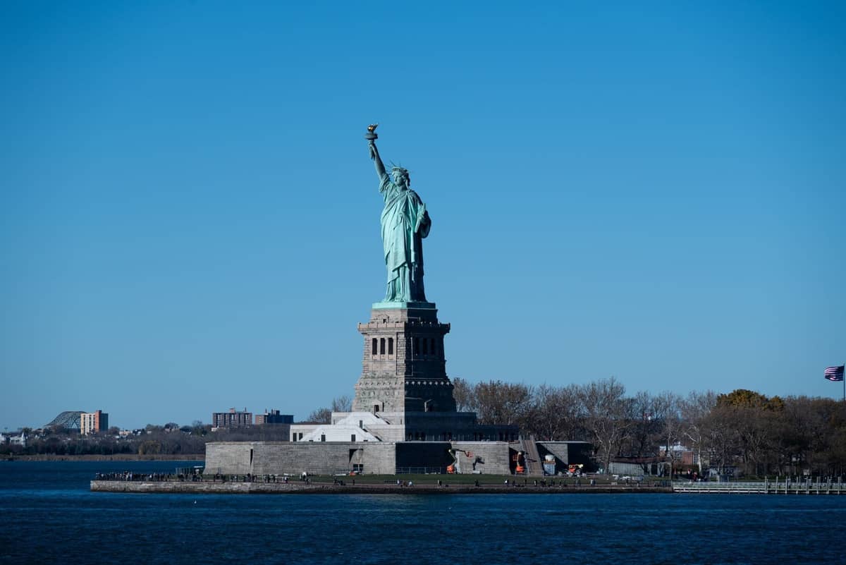 Weitwinkelansicht der Statue of Liberty auf ihrer Insel bei hellem Tageslicht, umgeben von Wasser, mit klarem blauem Himmel im Hintergrund.